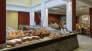 Zona de bufé con panes y pastelería variados, un cartel que indica opciones de desayuno, y vista del comedor al fondo.