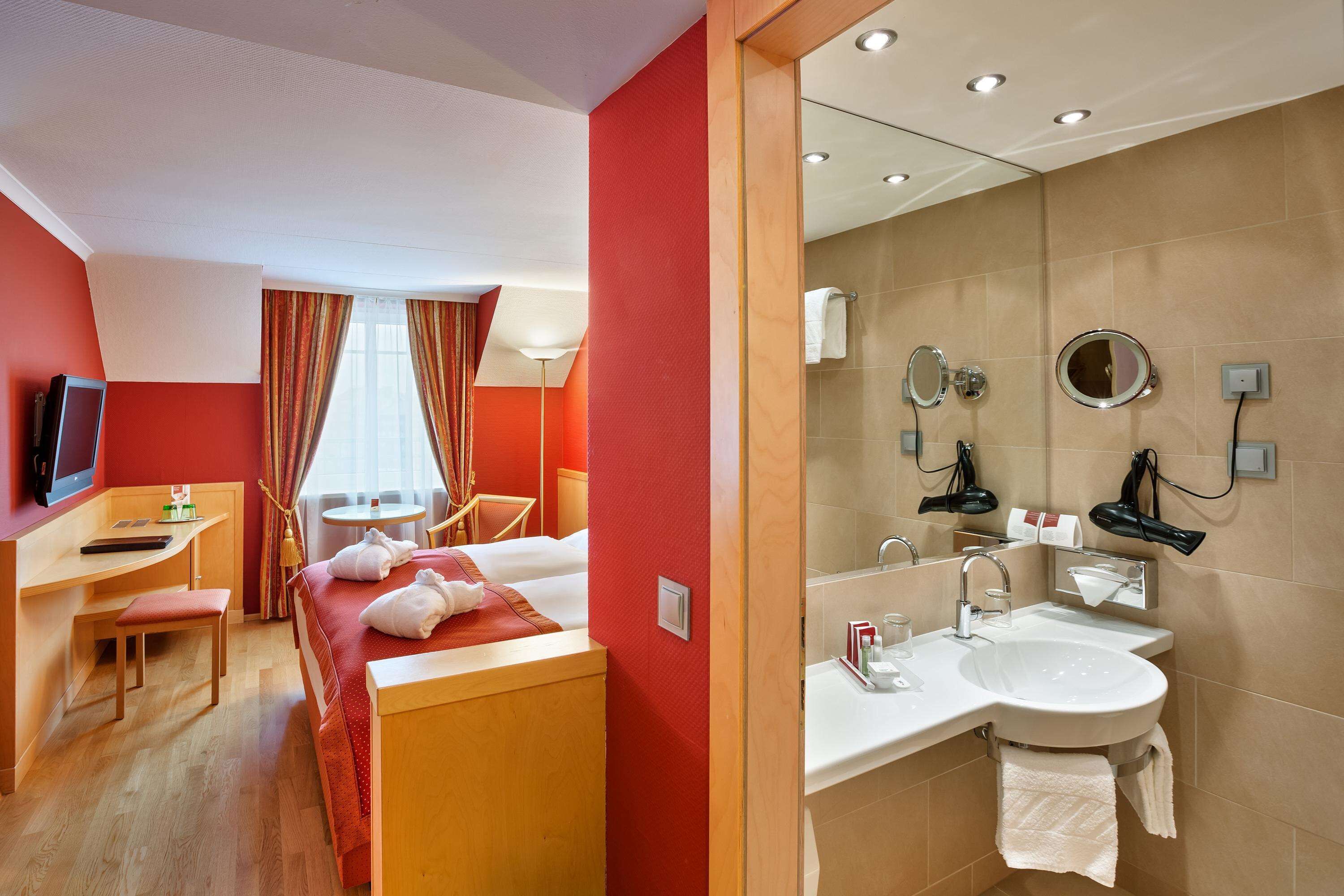 Quarto de hotel com cama, escrivaninha, cadeira, mesa pequena, TV e janela com cortinas. Inclui banheiro com pia, espelho e porta-toalhas.