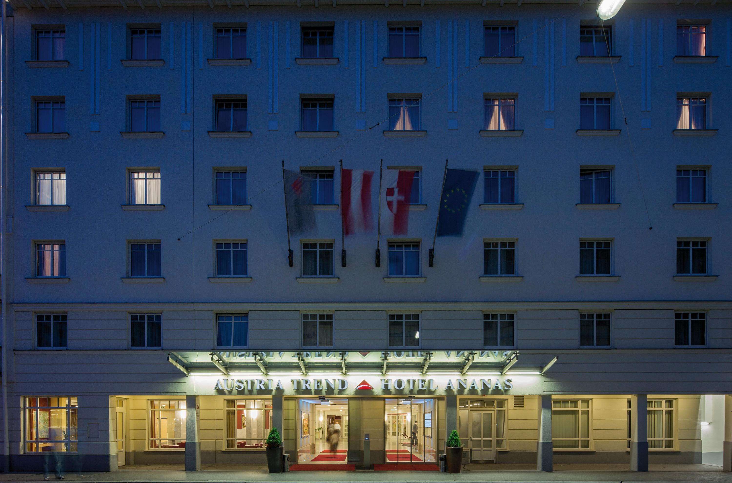 A hotel facade with flags, including the Swiss flag, and the name "AUSMIRIA TREND HOTEL ANATAS" illuminated.