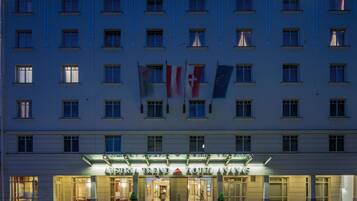 Fachada de hotel com bandeiras, incluindo a bandeira suíça, e o nome "AUSMIRIA TREND HOTEL ANATAS" iluminado.