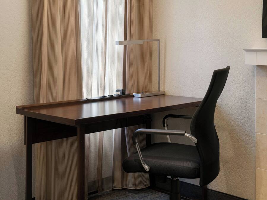 Quarto de hotel com uma escrivaninha de madeira, uma cadeira de escritório preta, um espelho e uma janela com cortinas leves.