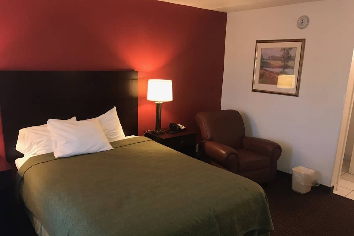 Hotellihuoneessa on sänky, tuoli, yöpöytä, lamppu ja seinällä oleva kehystetty kuva.