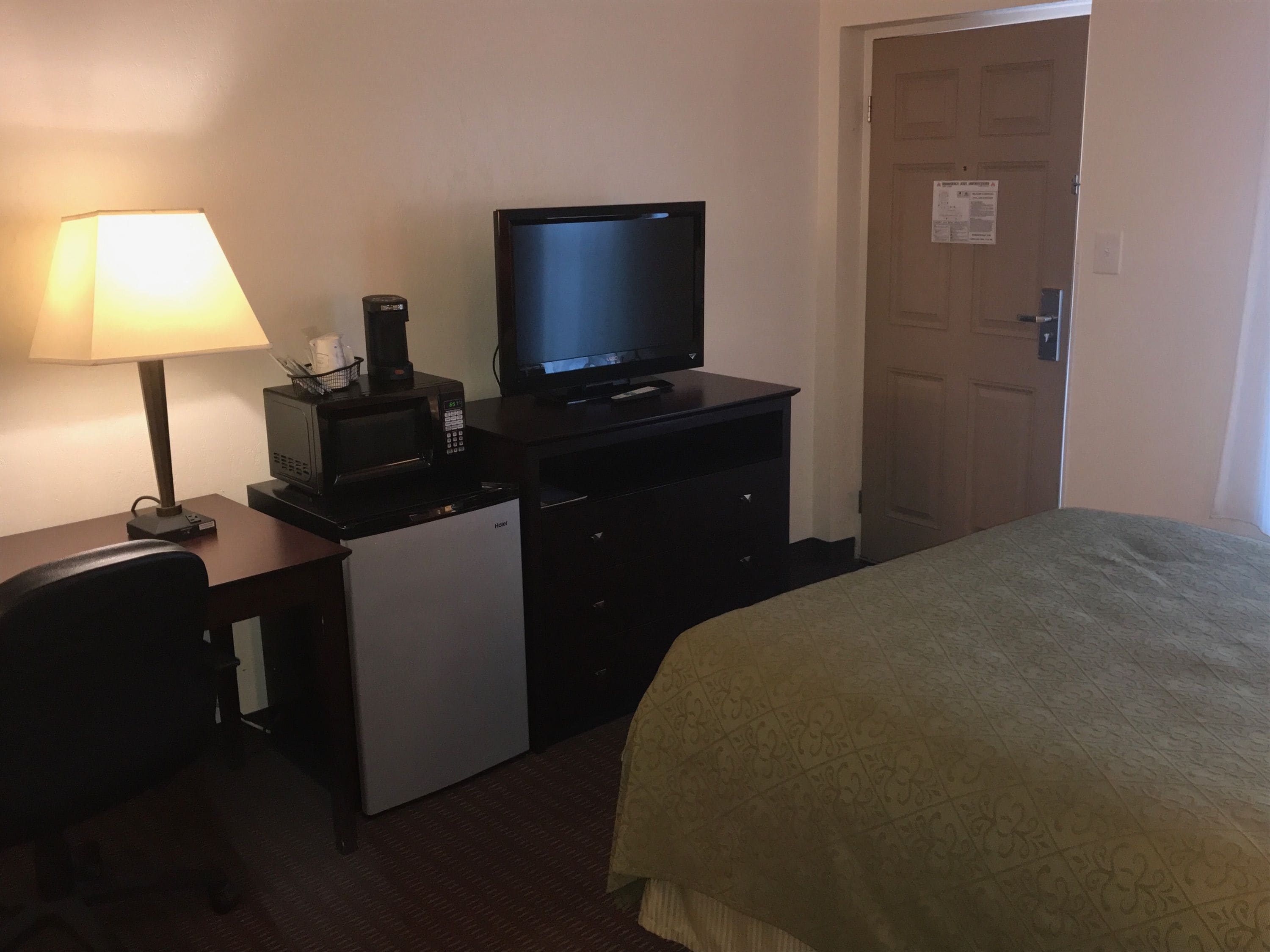 A hotel room with a bed, a desk with a lamp, a microwave, and a television.
