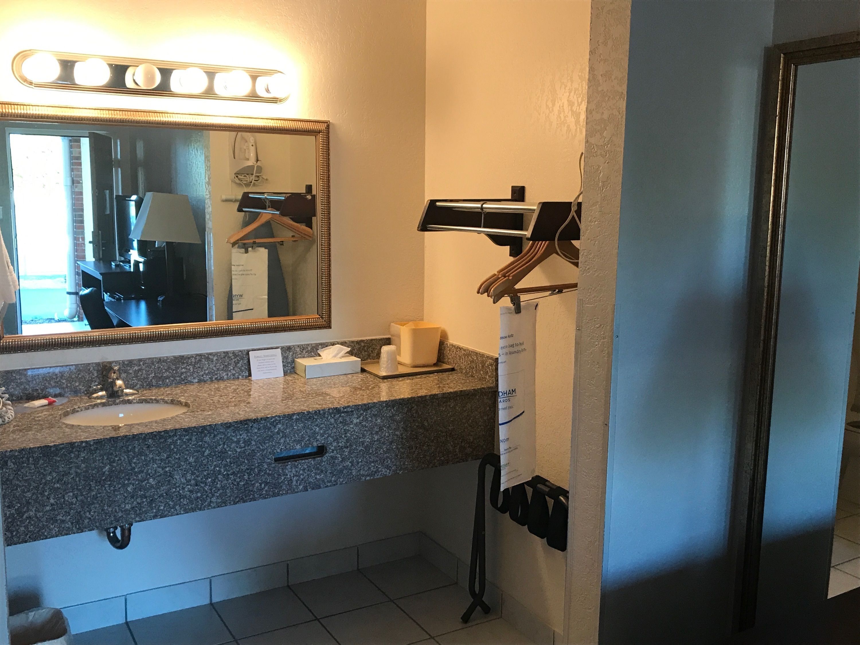 A bathroom with a granite countertop, a sink, a mirror with lights, a towel dispenser, and a paper towel dispenser.
