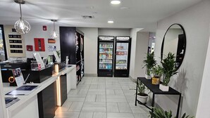 Zona de recepción con mostrador, dos refrigeradores llenos de bebidas, un espejo y plantas en macetas.
