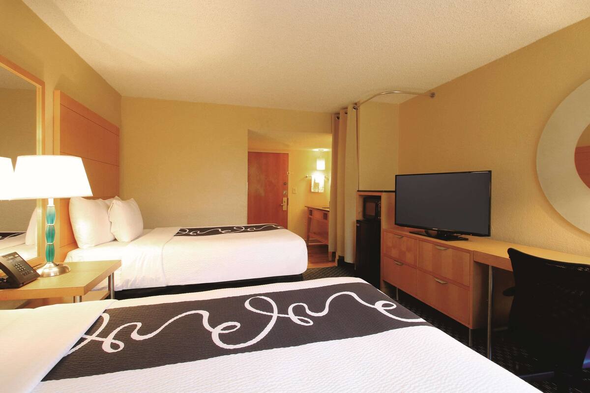 Una habitación de hotel con dos camas, un escritorio con televisor, y un baño visible al fondo.