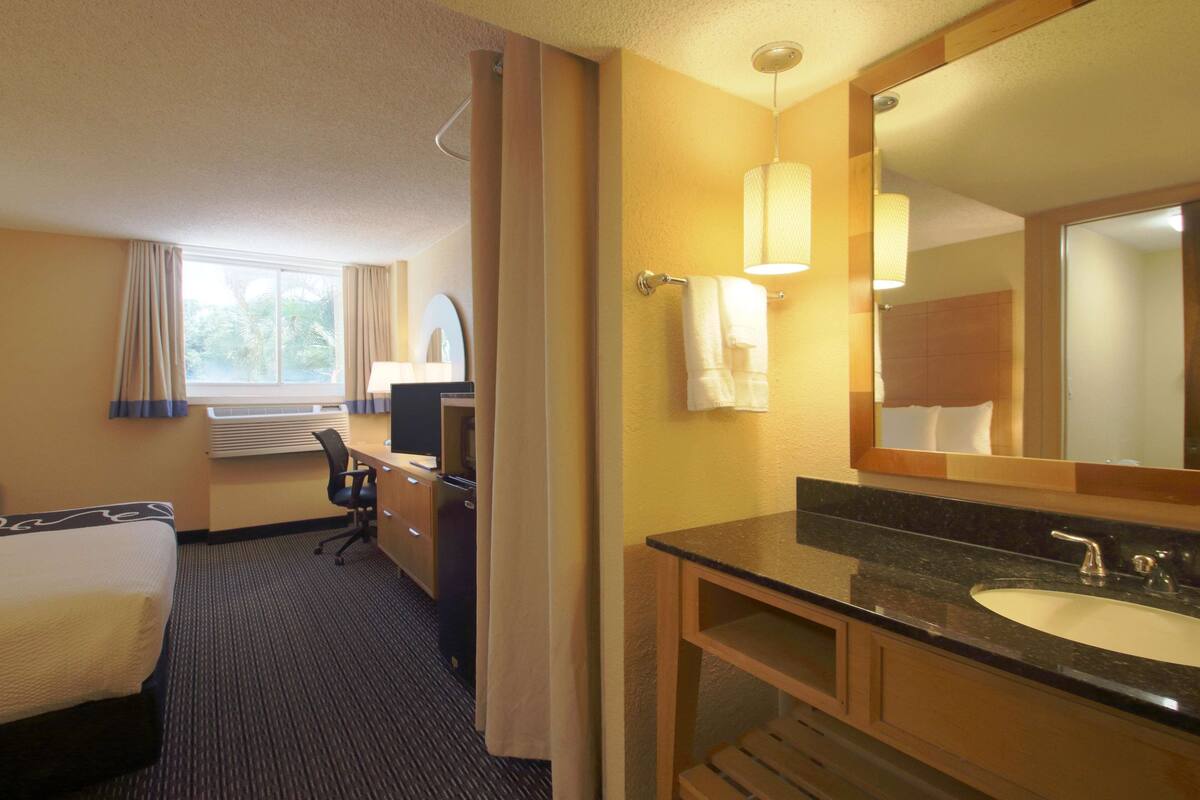 Una habitación de hotel con cama, escritorio, silla y baño con lavamanos y espejo.