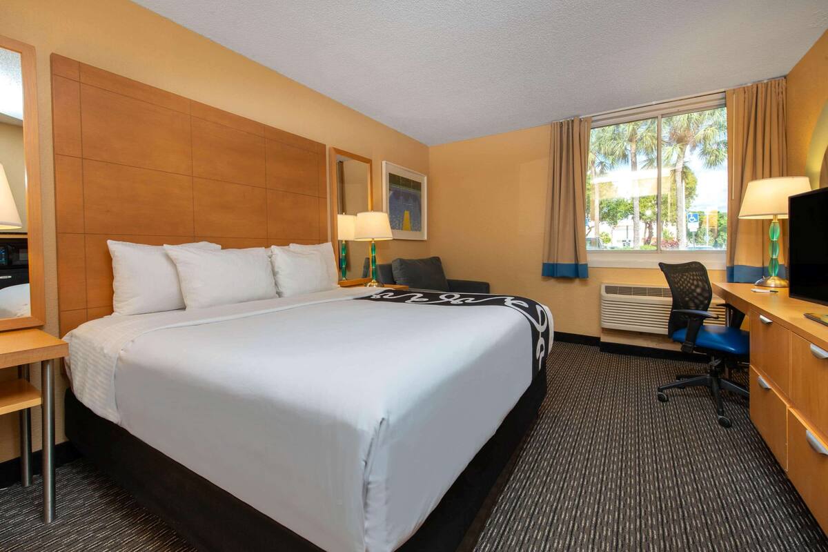 Habitación de hotel con una cama grande, un escritorio con computadora, una silla y una ventana con cortinas.