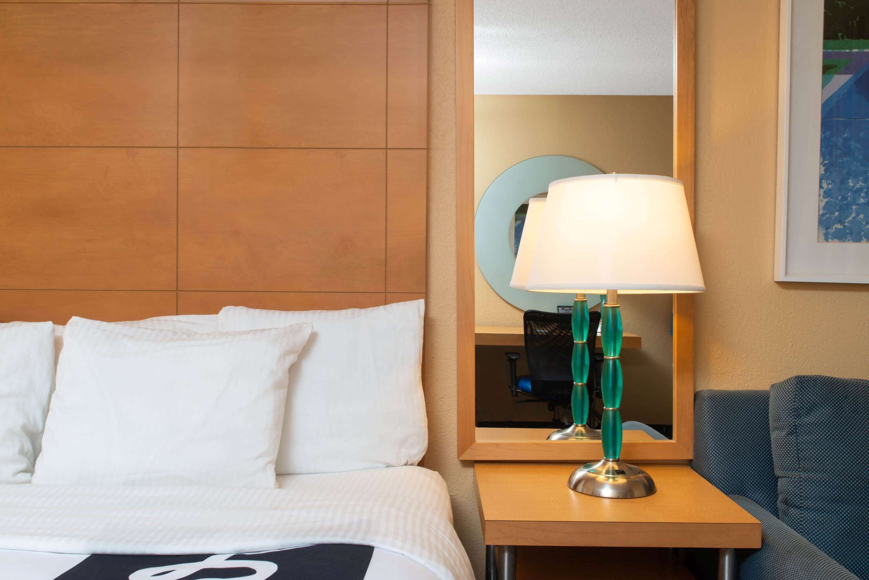 Habitación de hotel con cama, mesita de noche con lámpara, espejo y un cuadro en la pared.