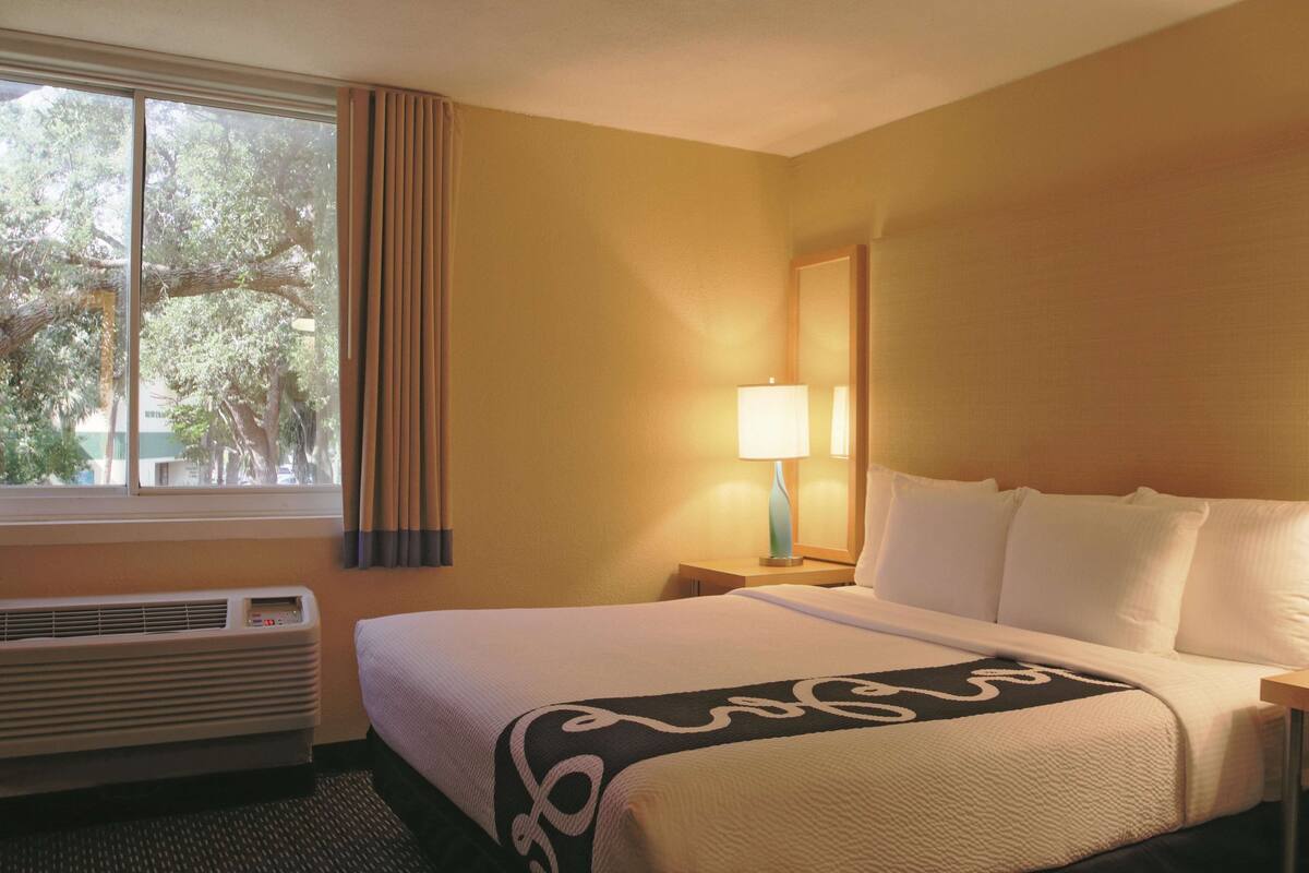 Una habitación de hotel con una cama grande, una ventana con cortinas, una lámpara de noche y un equipo de aire acondicionado.