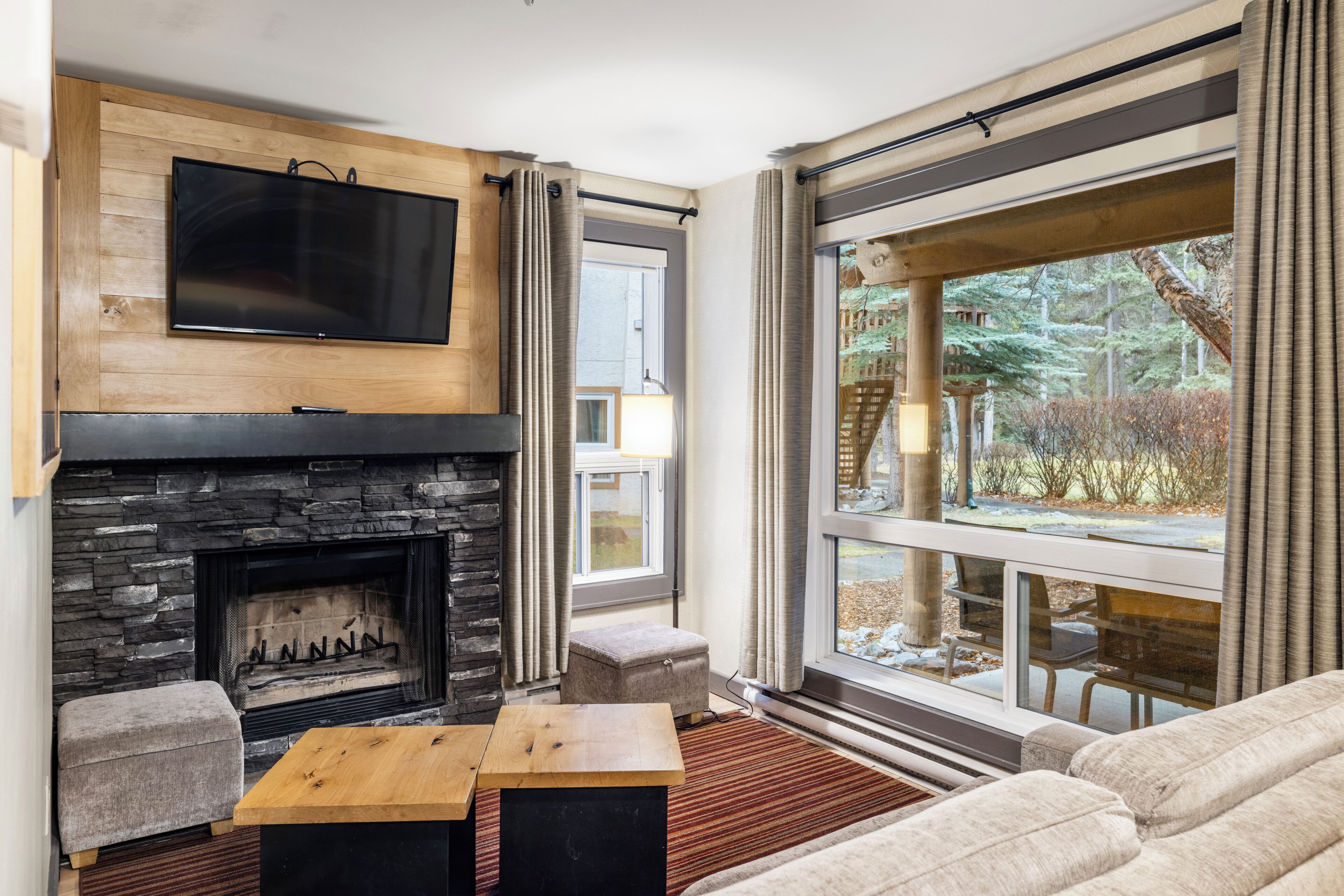 A modern living room with a stone fireplace, a flat-screen TV mounted above it, a large window with curtains, and a cozy seating area with a sofa and ottoman.