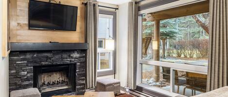 A modern living room with a stone fireplace, a flat-screen TV mounted above it, a large window with curtains, and a cozy seating area with a sofa and ottoman.