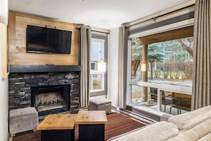 Una sala de estar moderna con chimenea de piedra, un televisor de pantalla plana montado arriba, un gran ventanal con cortinas y una zona de estar acogedora con sofá y reposapiés.