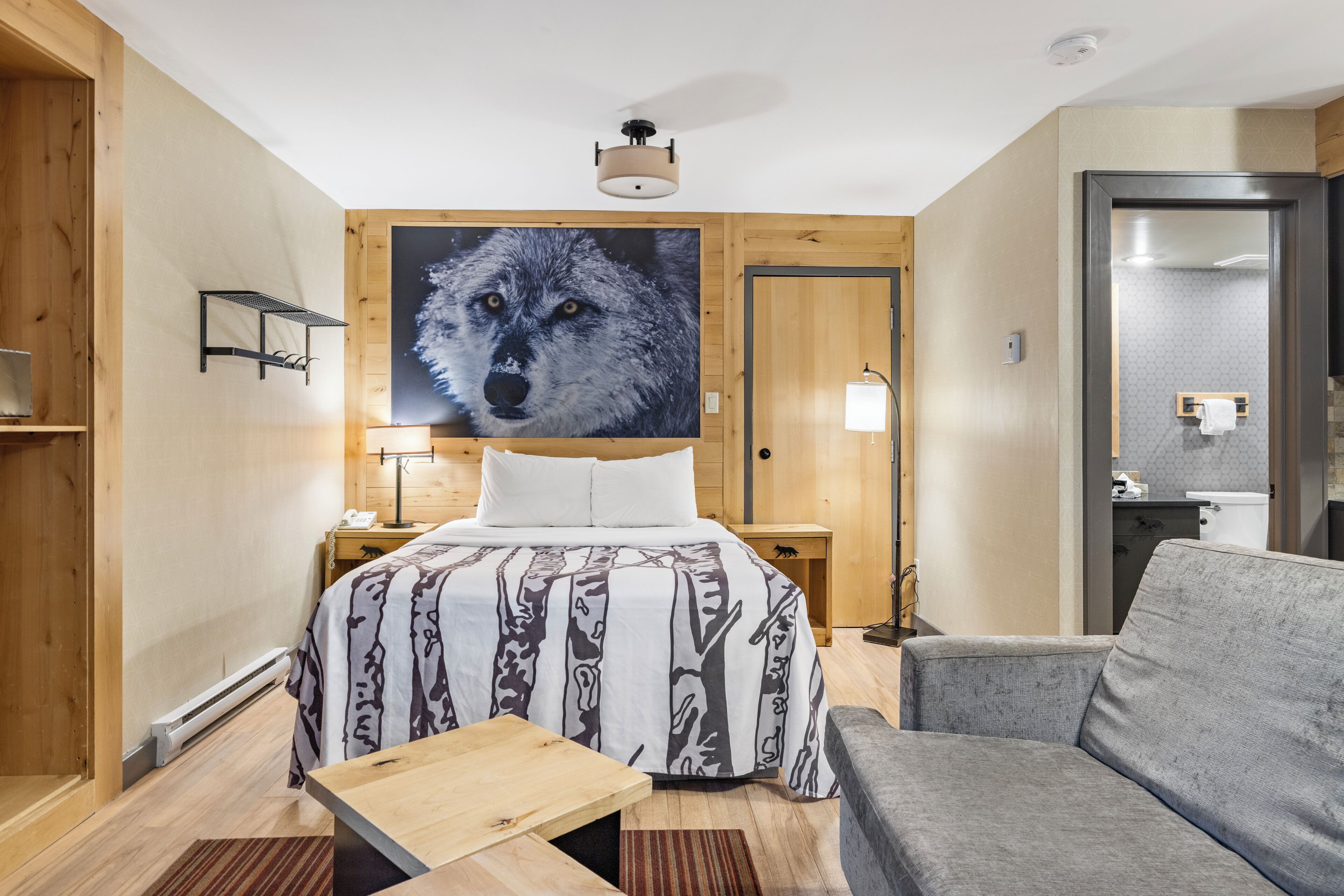 A hotel room with a bed, a sofa, a wooden nightstand, a wall-mounted lamp, and a large wolf portrait.