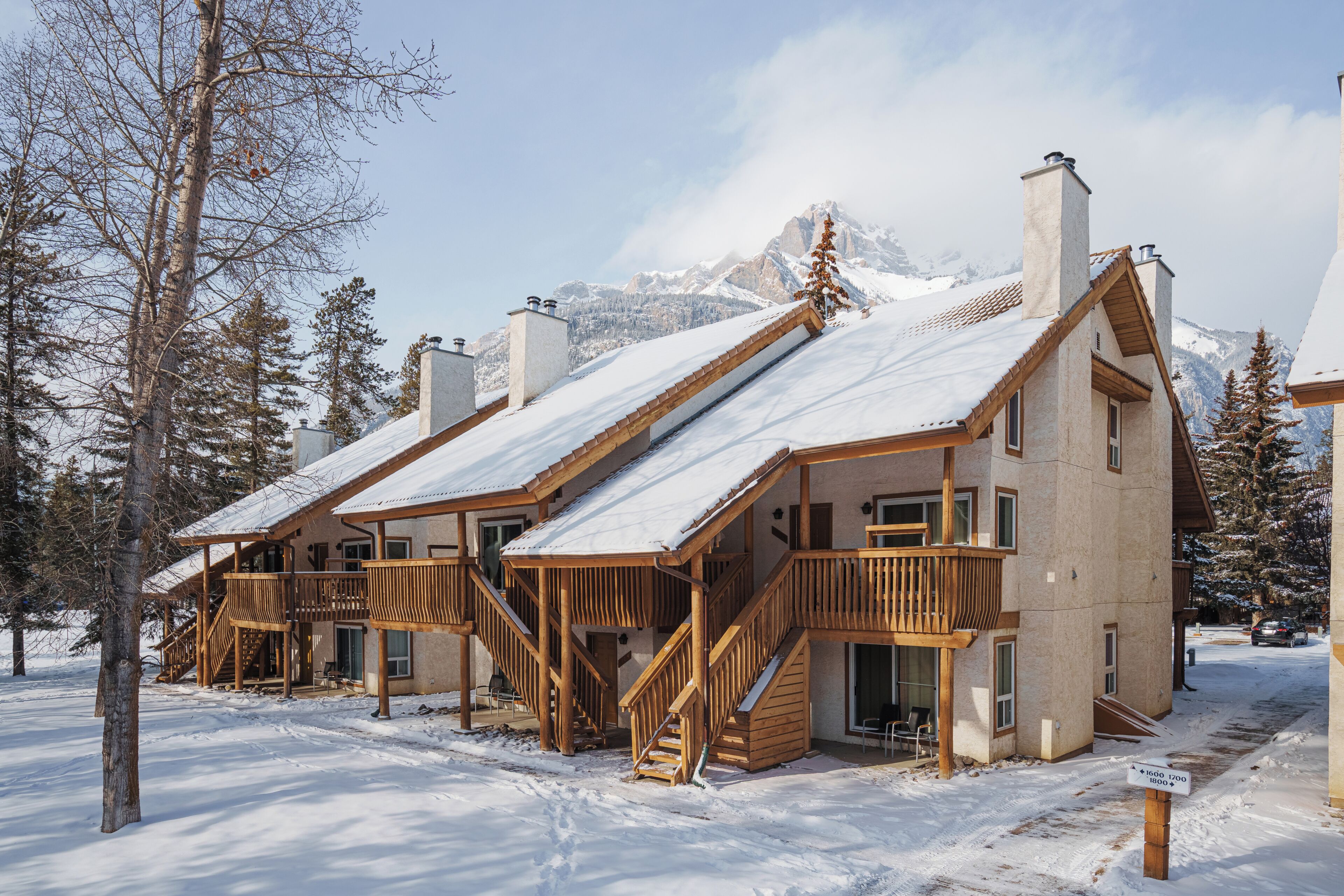 A chalet-style building with wooden balconies and a snow-covered roof, surrounded by trees and mountains.