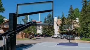 An outdoor basketball court with a clear blue sky, surrounded by residential buildings and parked cars.