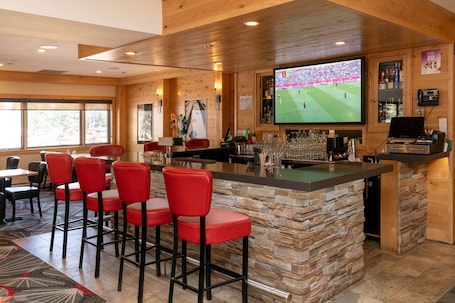 A bar area with a stone counter, red bar stools, and a large TV screen displaying a sports event.