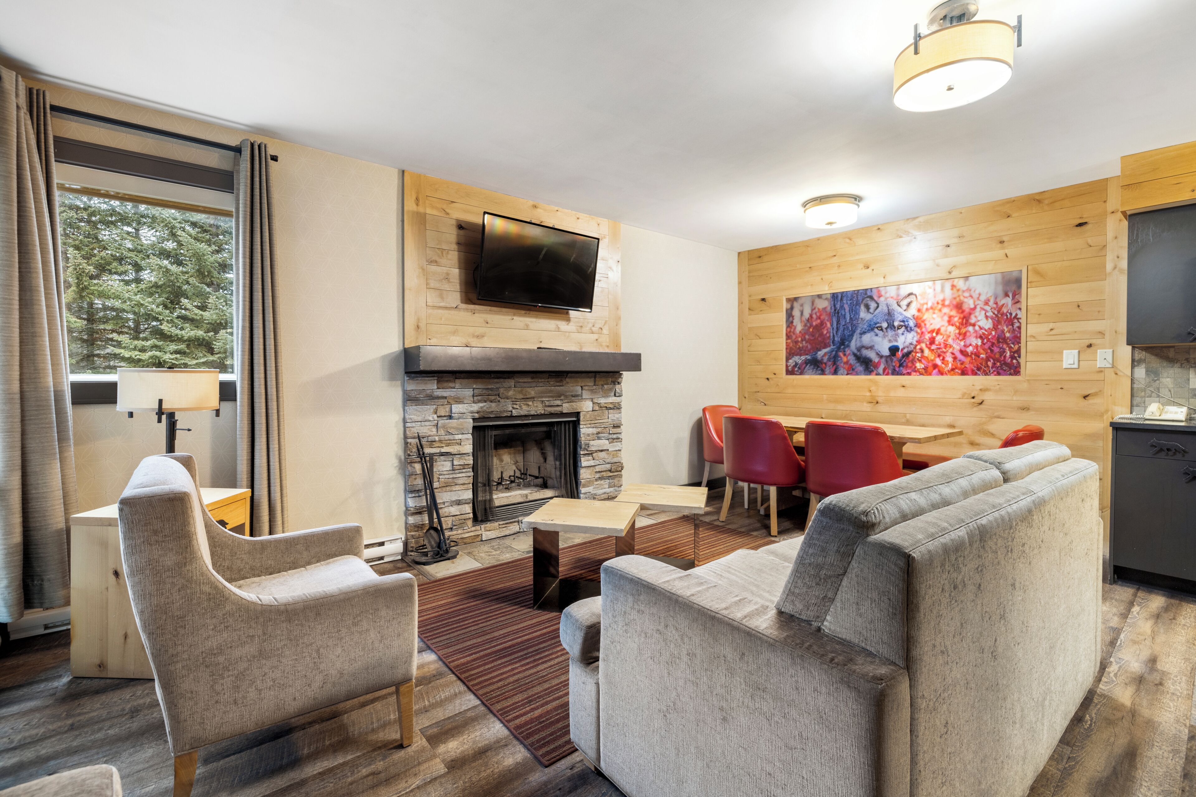 A modern living room with a stone fireplace, a flat-screen TV mounted above it, a dining area with red chairs, and a large painting on the wall.