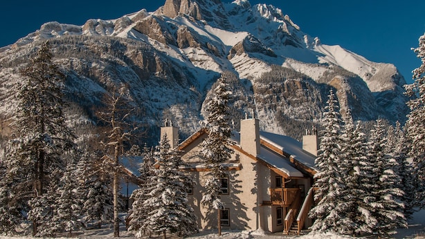 A mountain lodge with a wooden exterior, surrounded by snow-covered trees and a snow-covered mountain in the background.