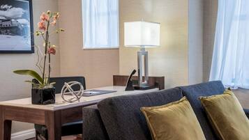 A modern hotel room with a dark grey sofa, two gold cushions, a wooden side table with a potted orchid, a framed photograph on the wall, and a bedside lamp.