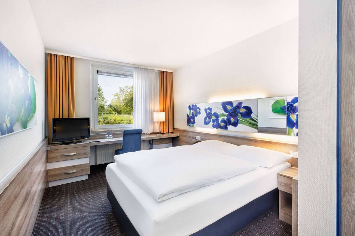 A hotel room with a bed, desk, chair, TV, and artwork.