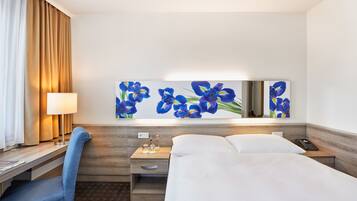 Ein Hotelzimmer mit einem Bett, einem Schreibtisch mit Lampe, einem Sessel, einem Nachttisch und einem Wandbild mit blauen Blumen.