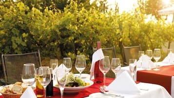 An outdoor dining setup with tables set for a meal, glasses, and a bottle of wine, surrounded by a vineyard.