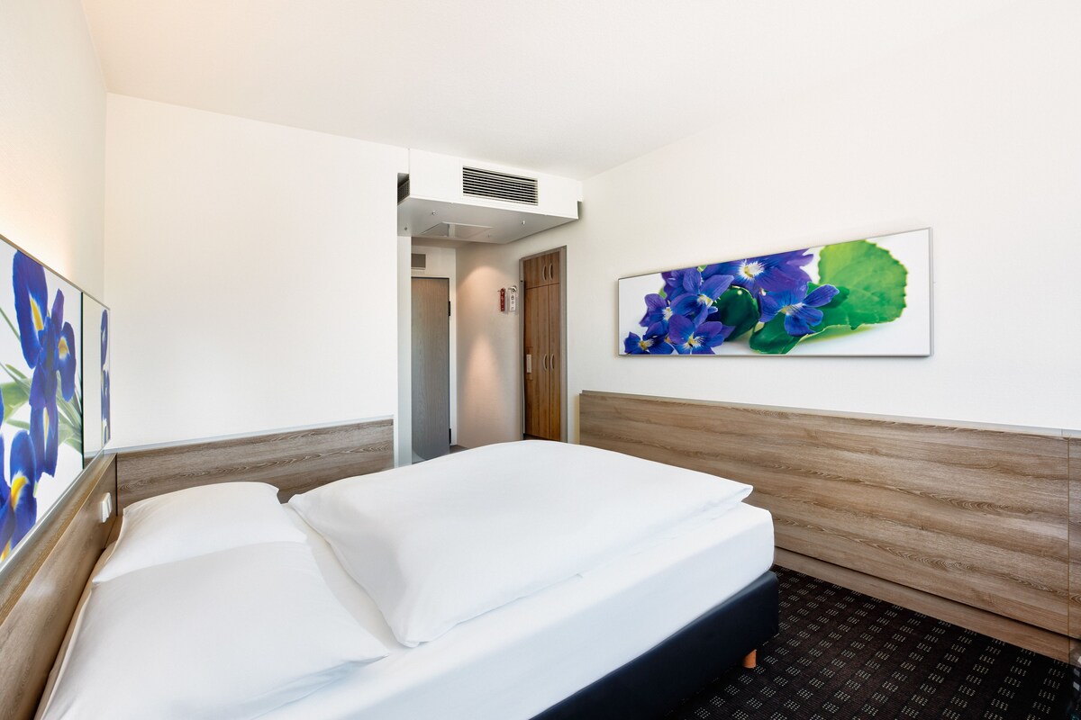 A hotel room with a bed, a wooden headboard, a wall-mounted air vent, and a framed floral artwork.