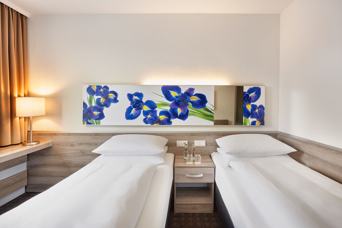 A hotel room with two single beds, a nightstand with a lamp, a wall-mounted mirror, and a floral artwork.