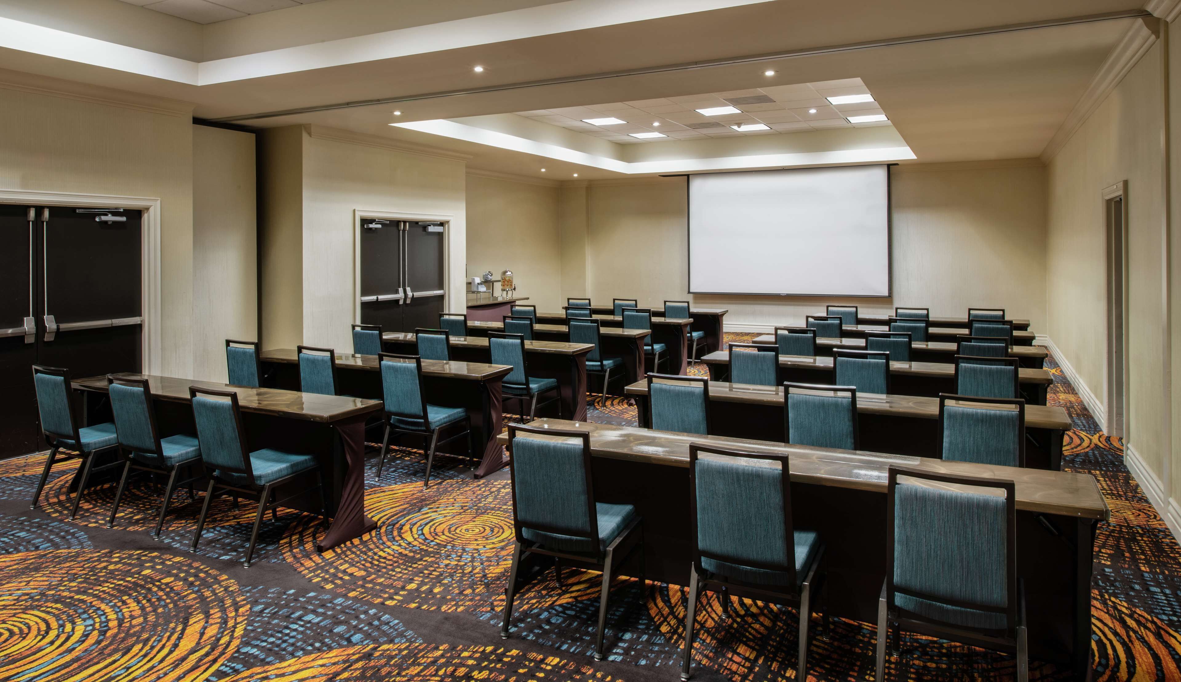 A conference room with rows of tables and chairs, a projector screen, and a carpeted floor.