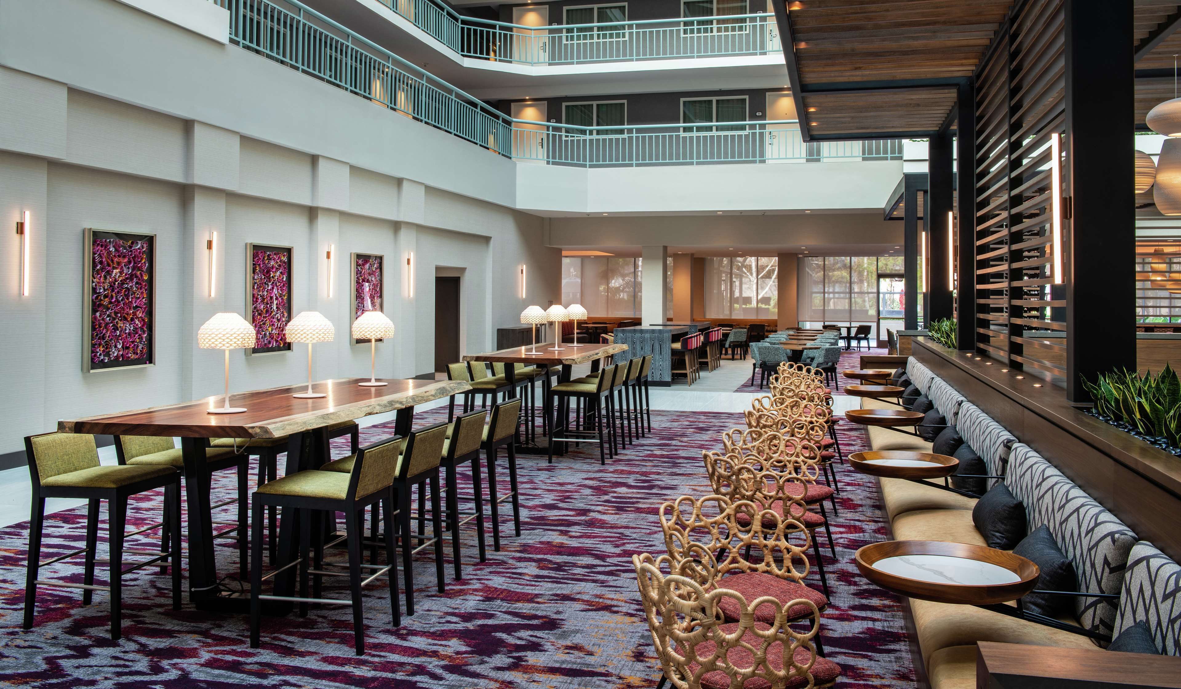 A spacious hotel lobby with a high ceiling, a central dining area with long tables and chairs, a seating area with sofas and round tables, and a staircase leading to an upper level.