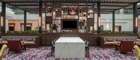 A spacious indoor area with a central seating area featuring a marble table and chairs, surrounded by shelves with decorative items and a large flat-screen TV.