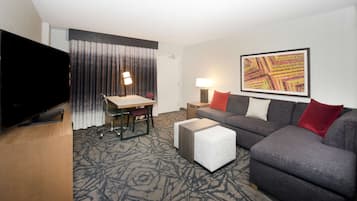 A hotel room with a flat-screen TV, a desk with a chair, a grey sofa, a small ottoman, a standing lamp, and a large abstract painting on the wall.