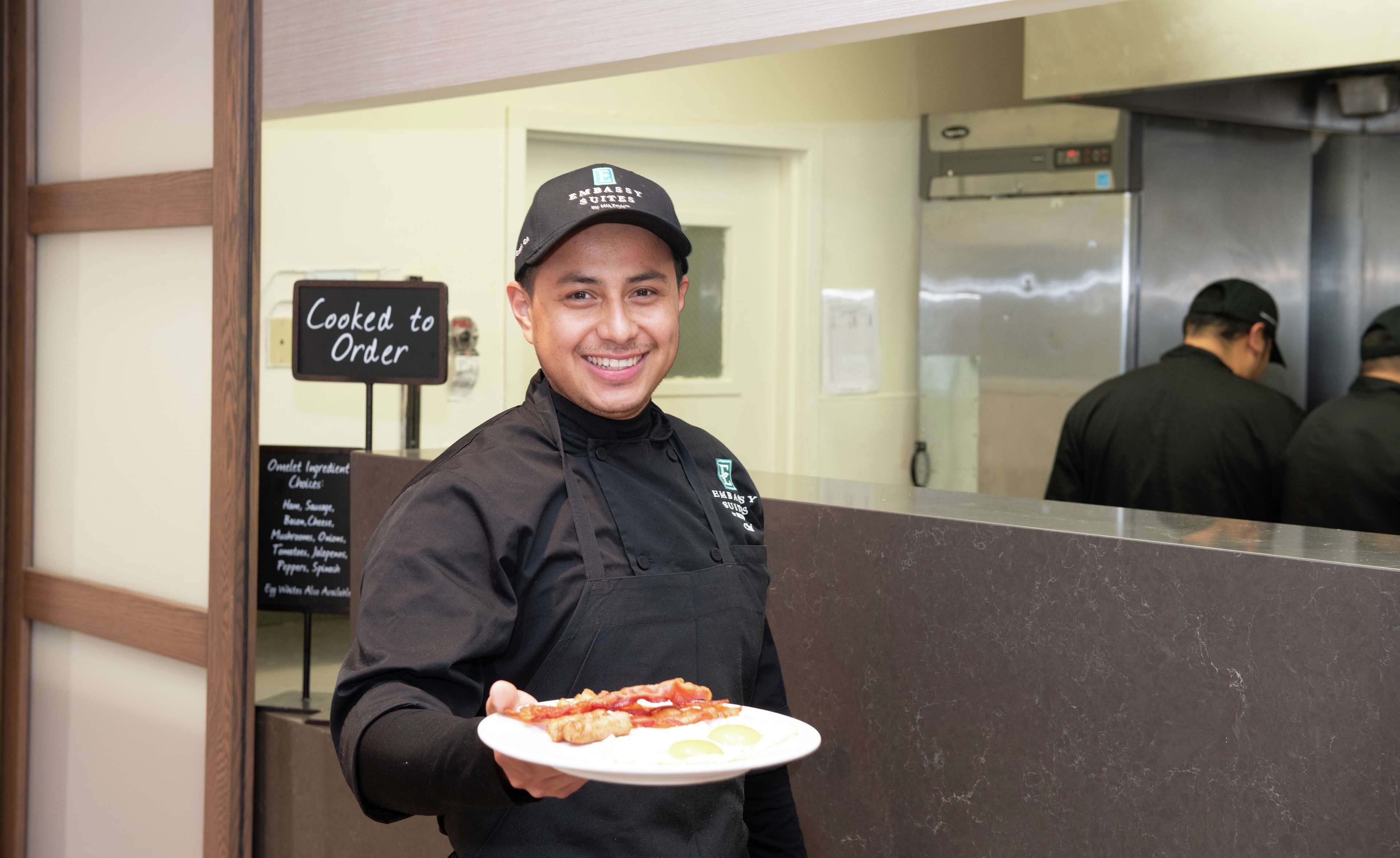 A person holding a plate of food in a kitchen area with a sign that reads "Cooked to Order".