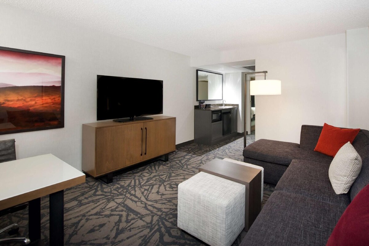 Habitación de hotel con televisión de pantalla plana, sofá, mesa de comedor, otomana pequeña, zona de cocina y ventana grande.