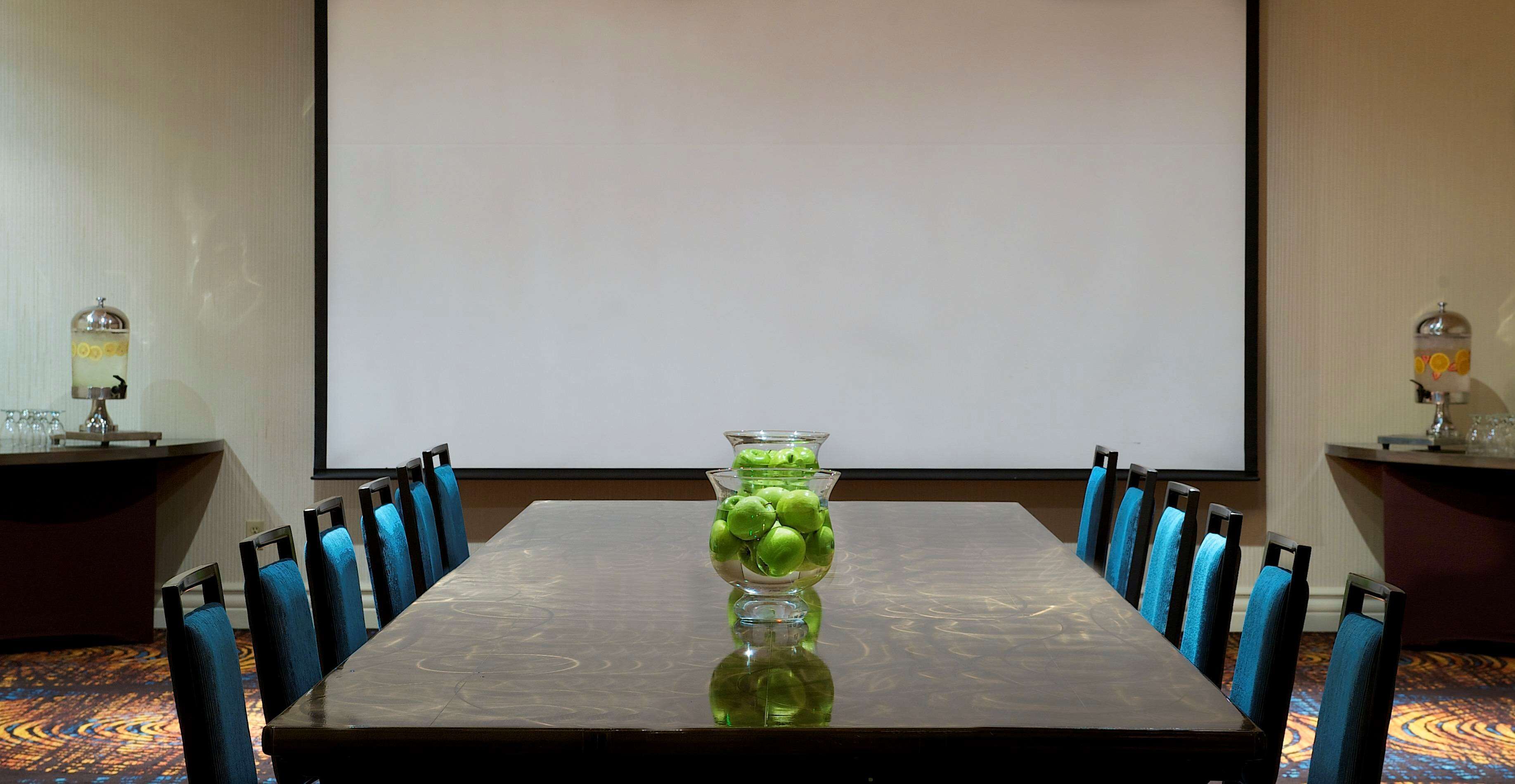 Un salón de conferencias con una larga mesa, sillas azules y una pantalla de proyector.