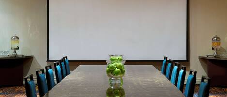 Un salón de conferencias con una larga mesa, sillas azules y una pantalla de proyector.