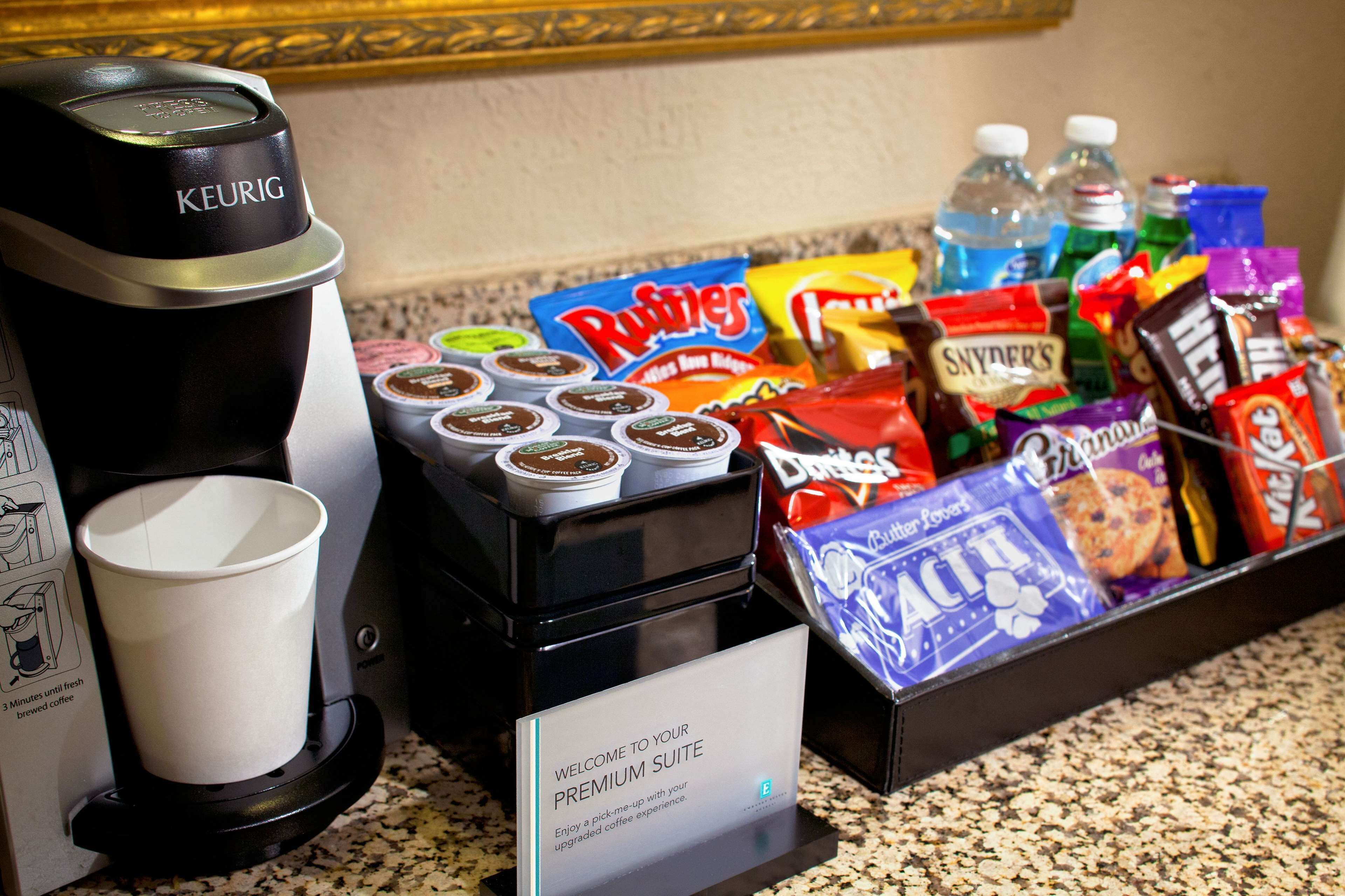 Una estación de café con una cafetera Keurig, vasos desechables y una variedad de bocadillos y bebidas.