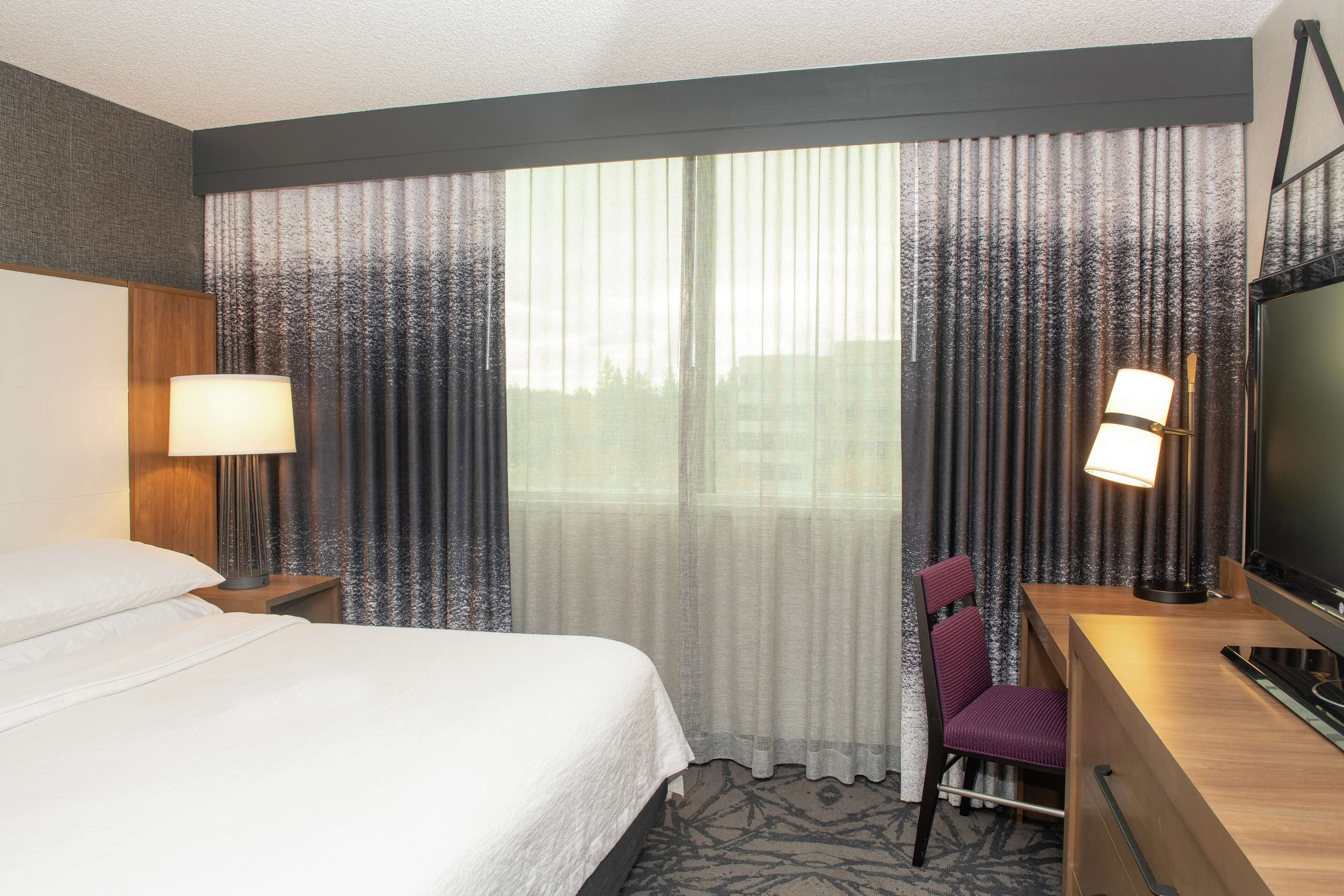 A hotel room with a bed, a desk with a chair, a TV, and two lamps.