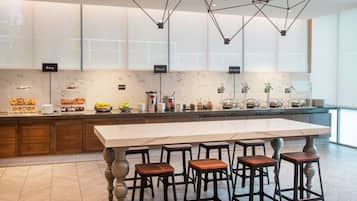 Une cuisine moderne dotée d'un îlot central, de tabourets de bar et d'un buffet pour le petit-déjeuner, avec des étiquettes indiquant les différents types de produits proposés : viennoiseries, porridge et plats chauds.