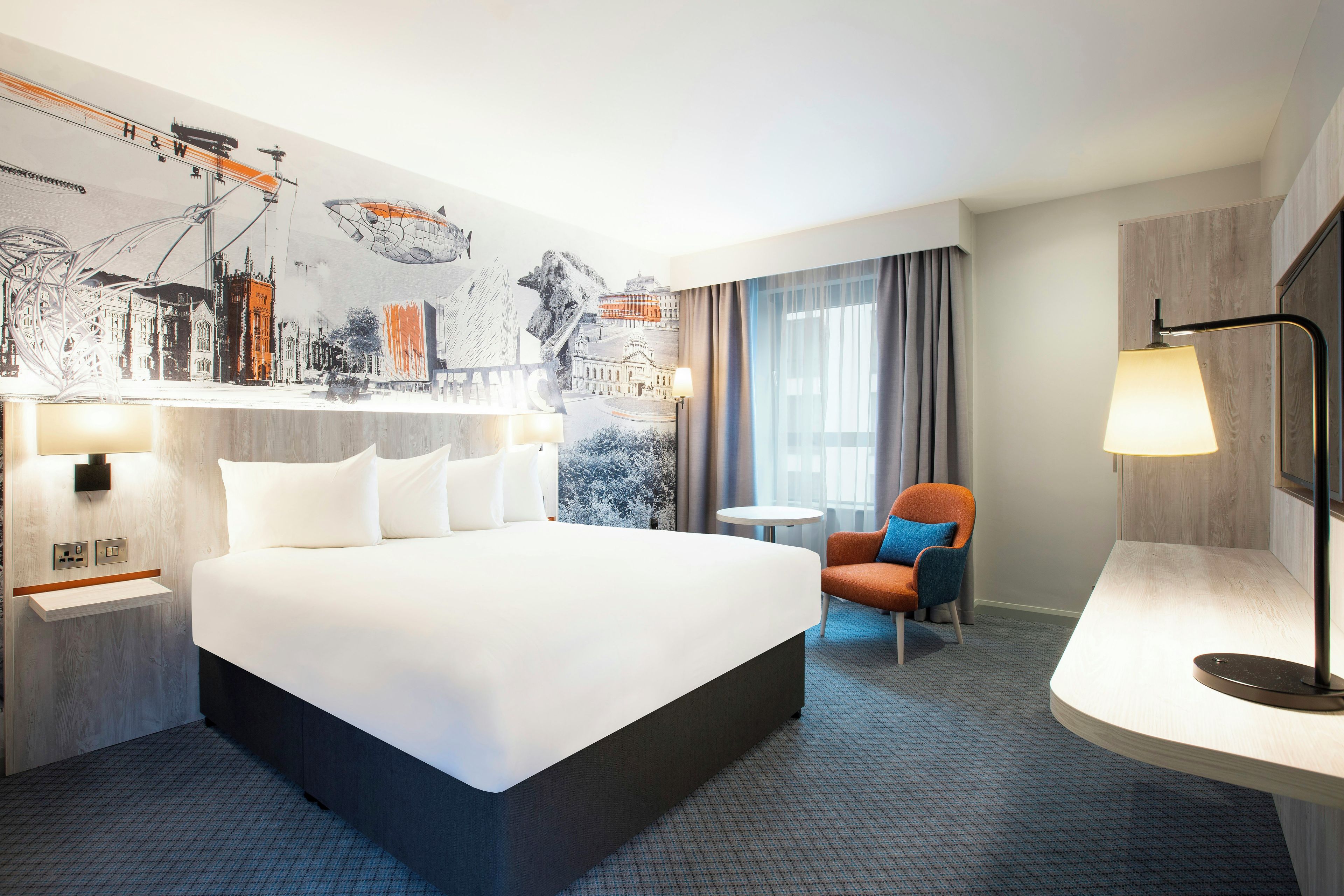 A hotel room with a large bed, a desk, a chair, a lamp, and a wall with a cityscape mural.