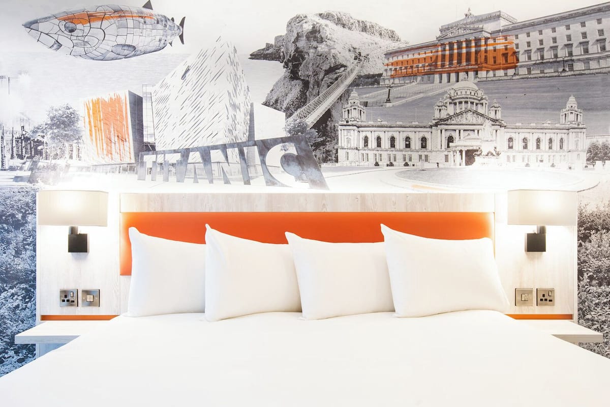 A modern hotel room with a large bed, orange headboard, and white pillows.