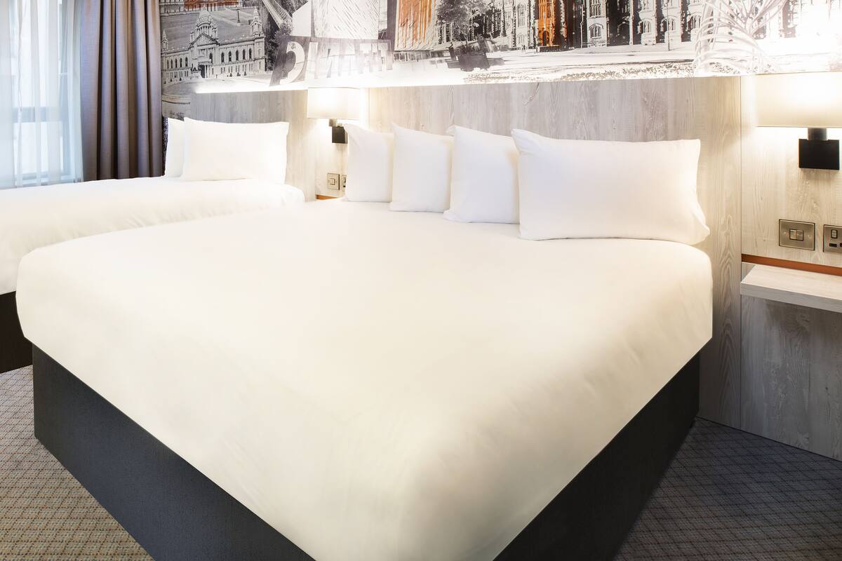 A hotel room with two large beds, a nightstand, and a wall-mounted lamp.