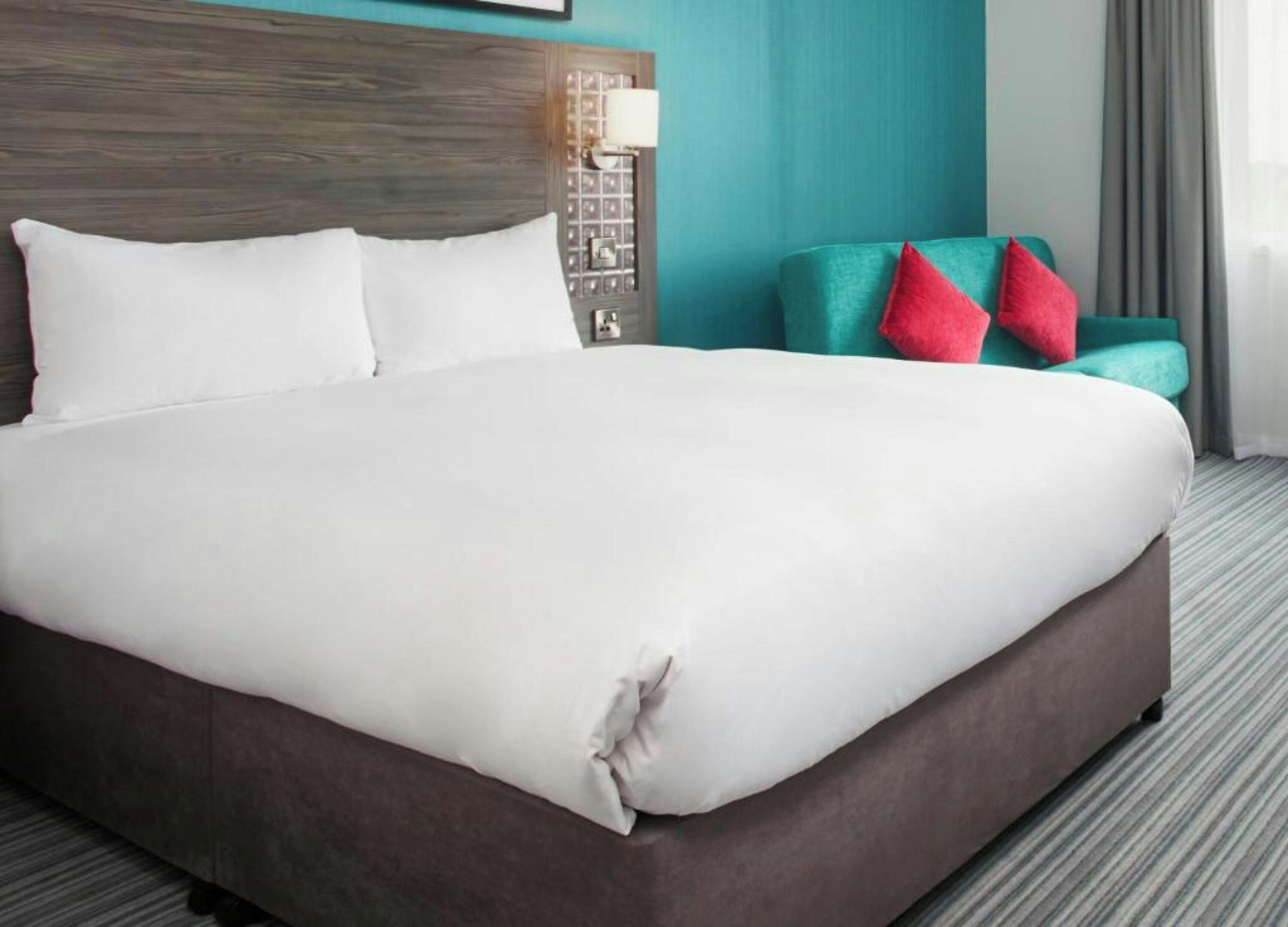 A large bed with white linens, a wooden headboard, a teal armchair with red pillows, and a wall-mounted lamp.