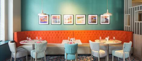 Ein Restaurant mit einem Türkis- und Orangeton, das eine lange, gepolsterte orangefarbene Bank, runde Tische mit Stühlen und gerahmte Bilder an der Wand zeigt.