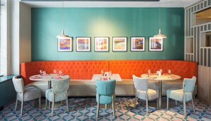 A restaurant with a teal and orange color scheme, featuring a long orange cushioned bench, round tables with chairs, and framed pictures on the wall.