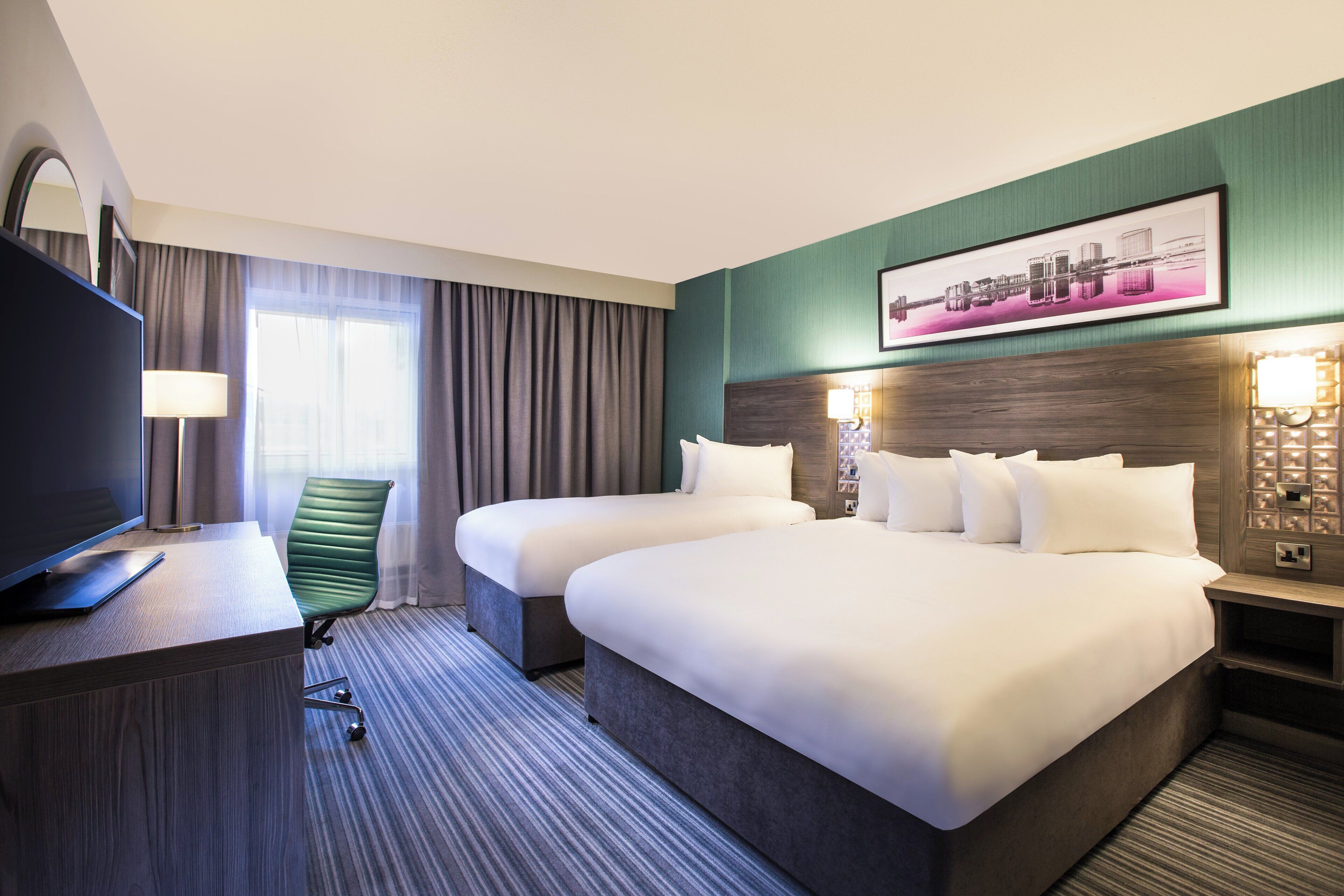 A hotel room with two beds, a desk with a TV, a chair, and a large window with curtains.