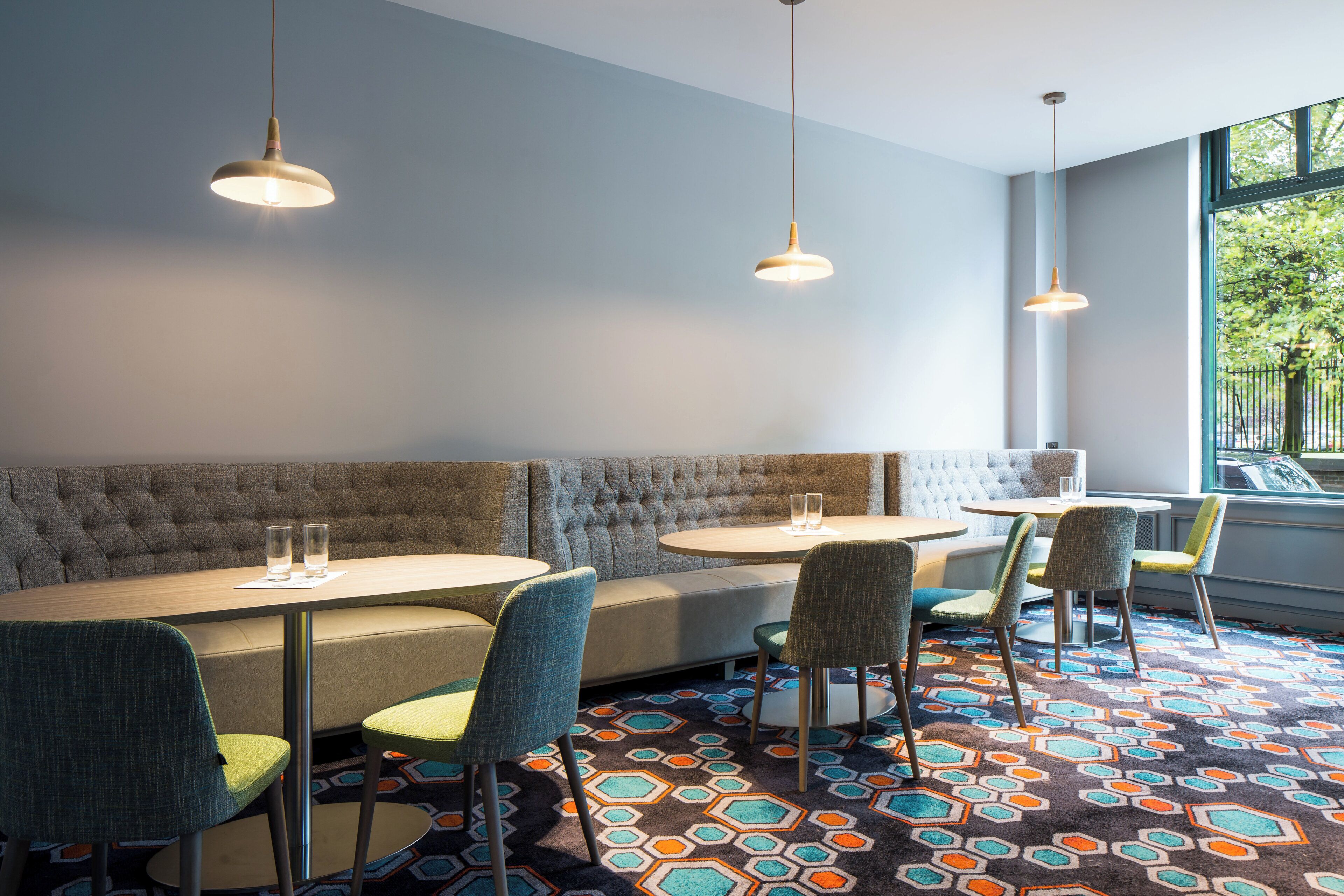 A modern dining area with a sofa, round tables, and chairs, featuring a geometric patterned carpet and pendant lighting.