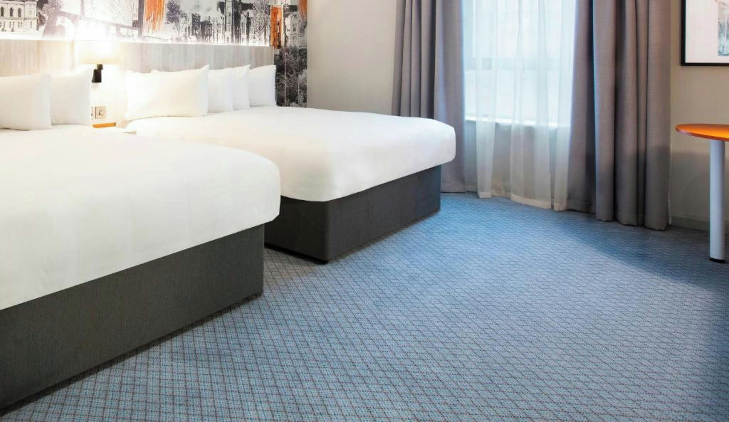 A hotel room with two beds, a small table, and a view of the city through the window.