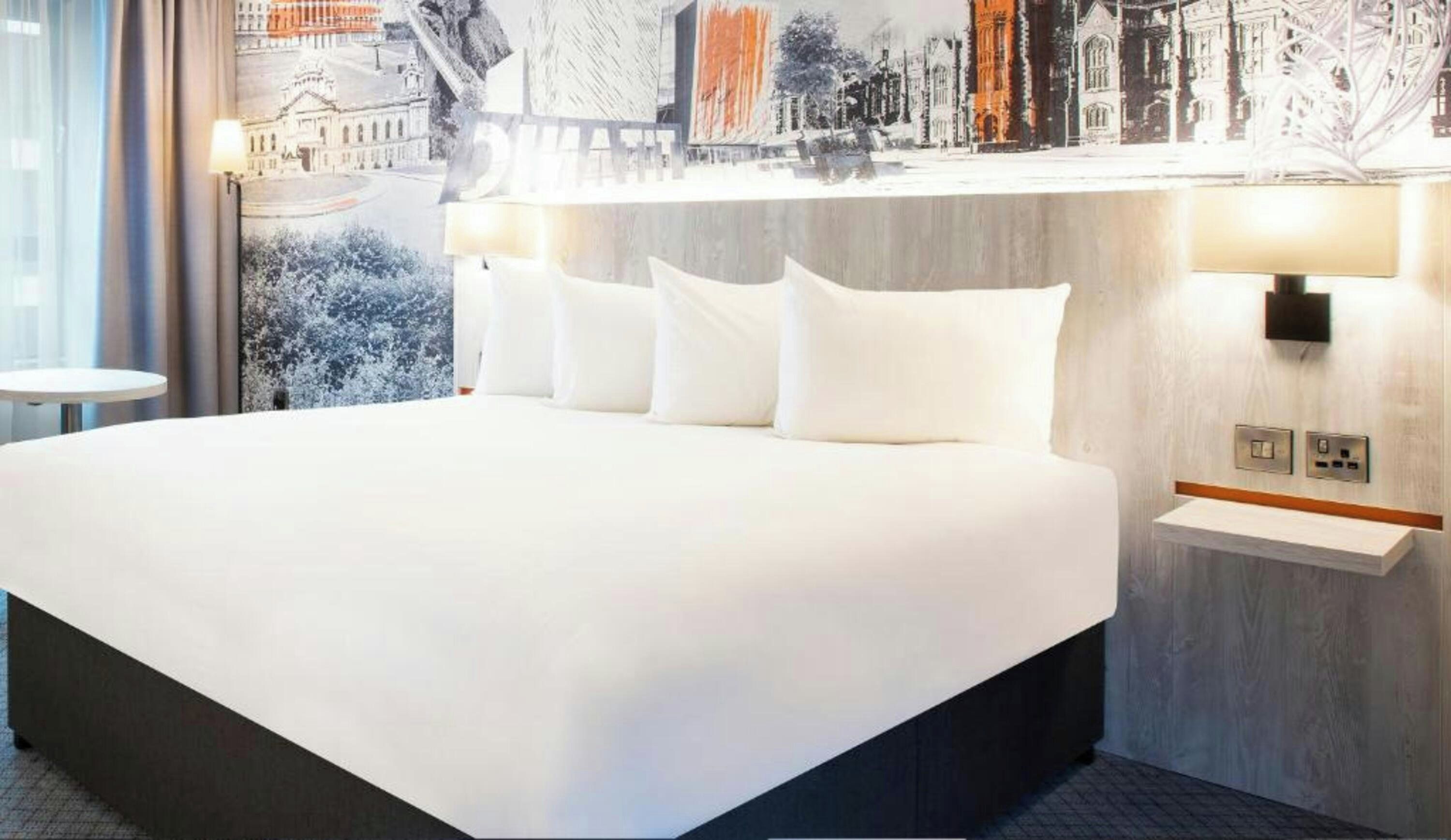 A hotel room with a large bed, a bedside table, a wall-mounted lamp, and a view of a cityscape through the window.